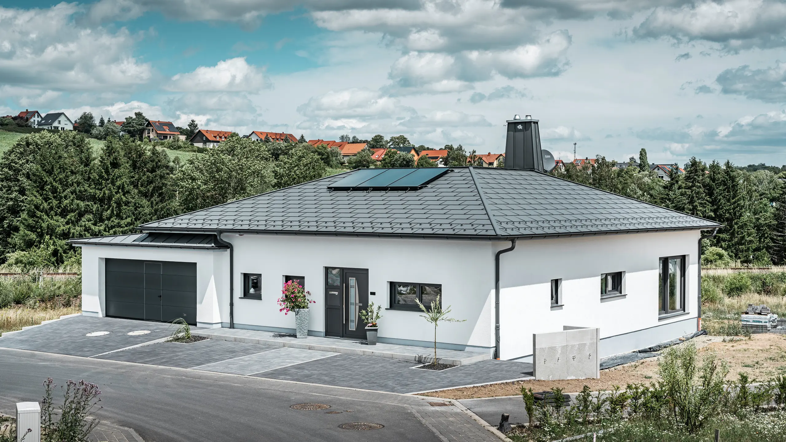Elegantna samostojeća kuća u Ilmenauu, Turingija, s visokokvalitetnim PREFA krovna ploča u boji P.10 tamnosiva, opremljena robusnim R.16 krovnim pločama. Garaža je u boji i stilu usklađena zahvaljujući upotrebi PREFALZ-a. Upadljive značajke nekretnine su funkcionalno dizajnirani atrij i dimnjak ukrašen PREFA bikom, koji naglašavaju moderan dizajn i inovativnu krovnu tehnologiju tvrtke PREFA. Solarni paneli besprijekorno se uklapaju u održivi koncept kuće. Dobro promišljen cjelokupni koncept bungalova zaokružuje PREFA sustav za odvodnju krova, koji je također u tamnosivoj boji.