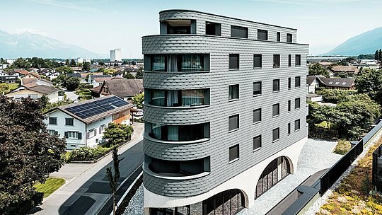 Zračni prikaz Bernerhausa u Buchsu s upečatljivom, trokutastom kubaturom i nježno zaobljenim balkonima. Fasada od PREFA zidnih šindra u P.10 svijetlosivoj boji obavija zgradu poput ljuskaste aluminijske kože i stvara živopisnu igru svjetla i sjene. Iznad masivnog betonskog postolja s elegantnim lukovima, lagana, otporna na vremenske uvjete PREFA aluminijska fasada djeluje posebno precizno, dugotrajno i gotovo bezdržavno – moderan vizualni naglasak usred alpske gradske panorame.