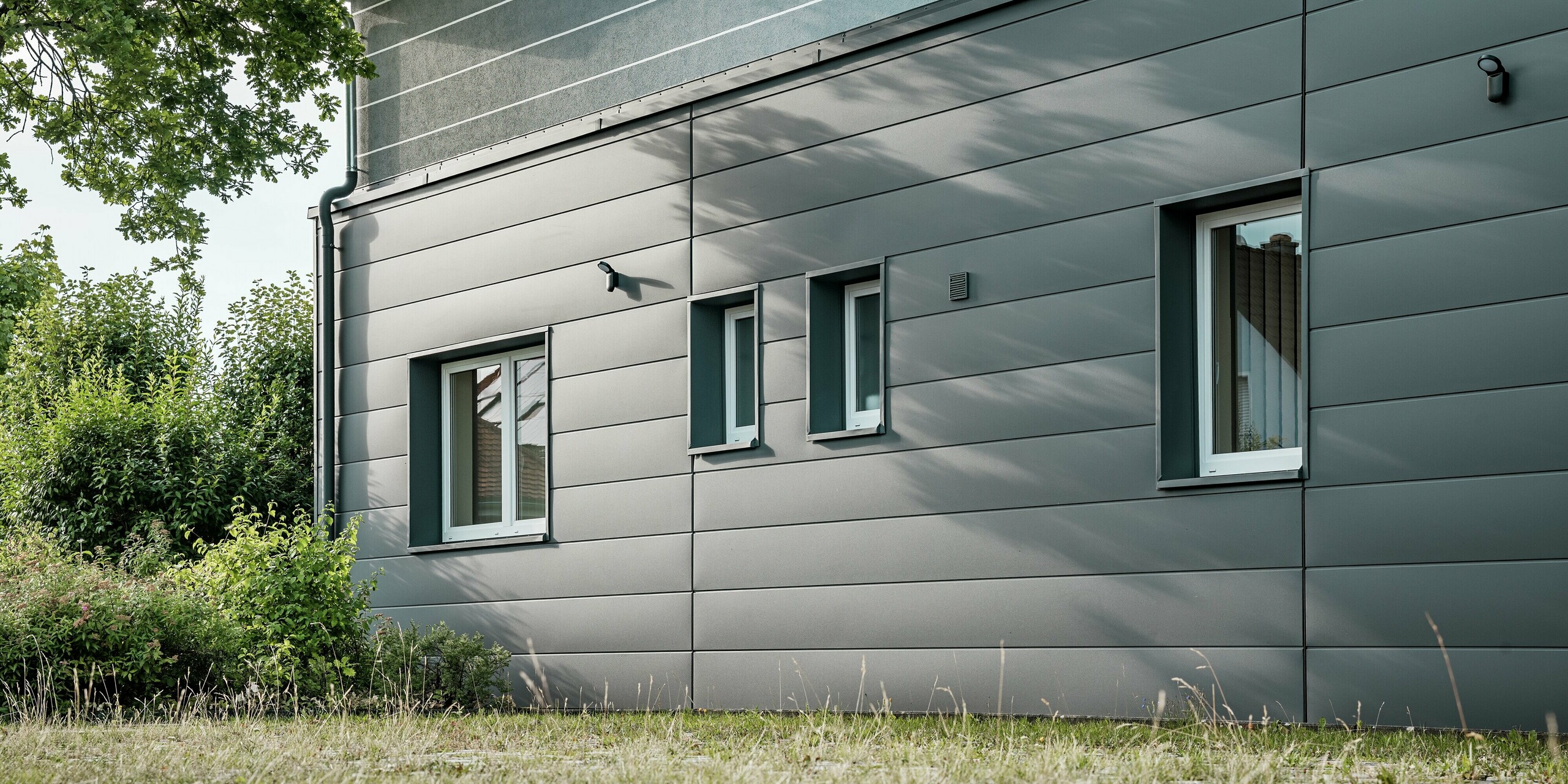 Das Bild zeigt einen Detailausschnitt der modernisierten Fassade der Kaminkehrerinnung in Bindlach, die mit PREFA Sidings in der eleganten Farbe P.10 Dunkelgrau ausgestattet wurde. Die klar strukturierten Aluminium-Paneele verleihen dem Gebäude ein professionelles und zugleich ästhetisches Erscheinungsbild. Besonders auffällig sind die harmonischen Schatten, die das Spiel von Licht und Architektur betonen. Dank der widerstandsfähigen und pflegeleichten Eigenschaften der PREFA Sidings bleibt die Fassade auch langfristig in einem hervorragenden Zustand.