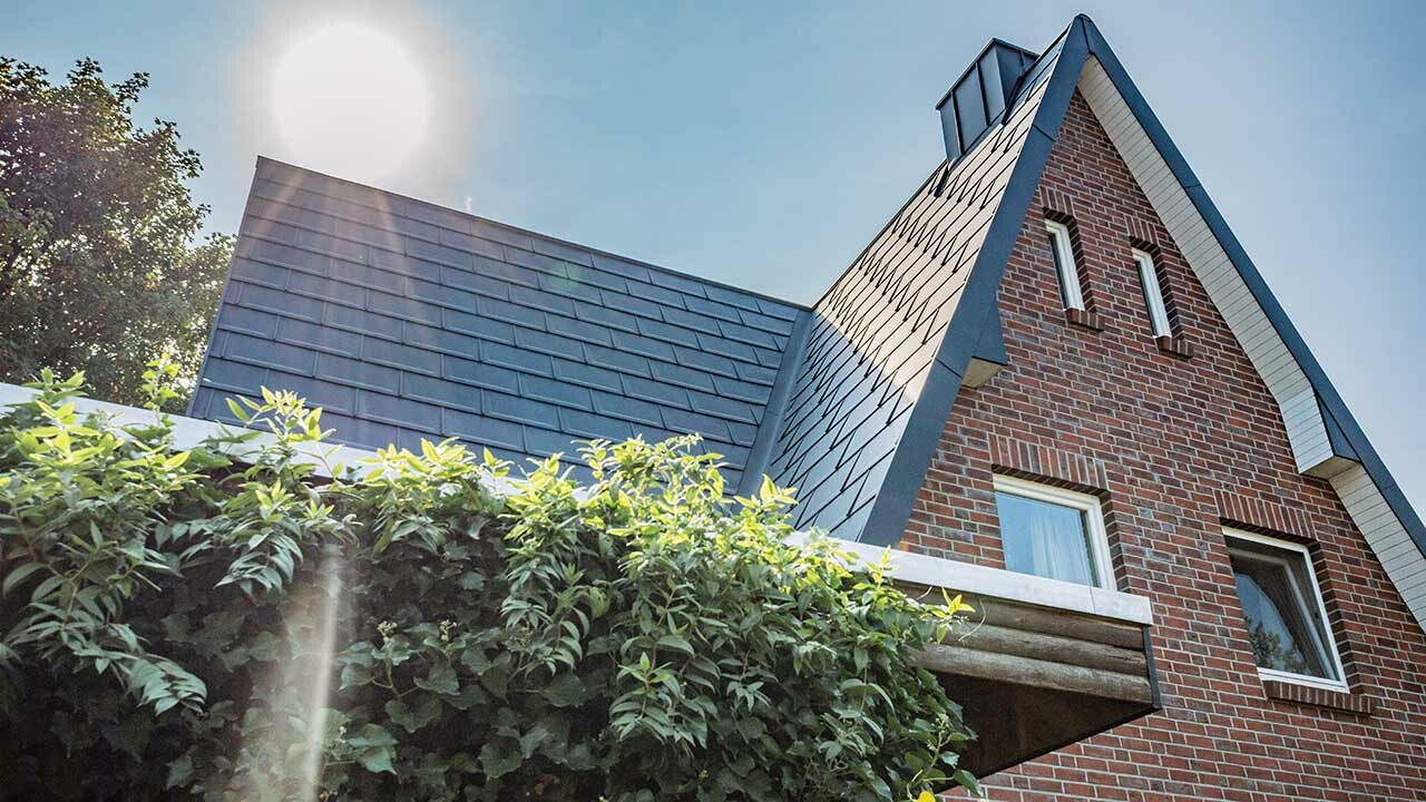 Dvostrešni krov pokriven jednostavnom PREFA aluminijskom krovnom pločom R.16 u antracit boji. Fasada je rustikalna klinker fasada, iza kuće upravo izlazi sunce.