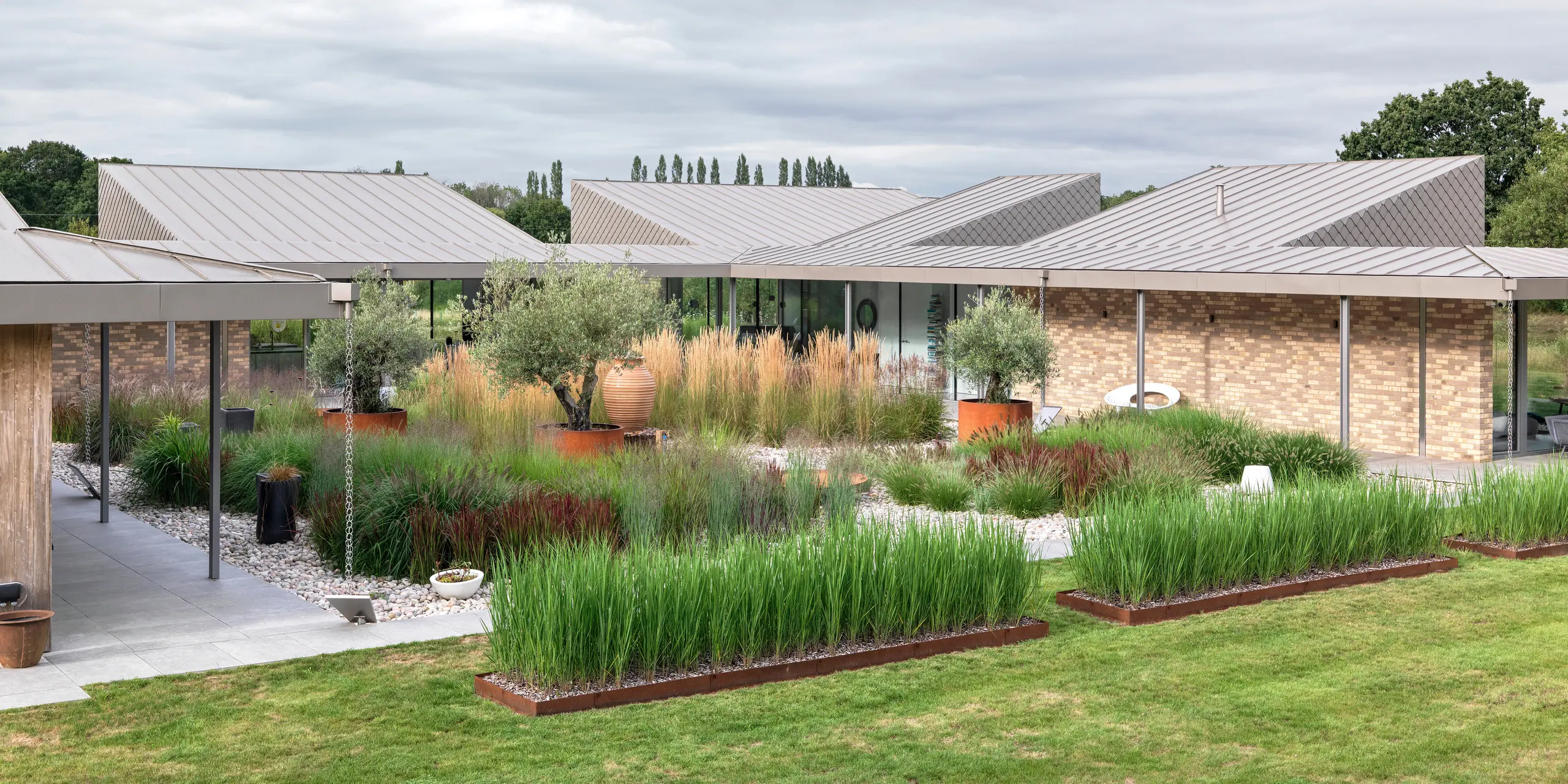 Großzügige Gartenansicht eines modernen Bungalows in West Sussex mit mehrfach gegliederten PREFALZ Aluminiumdächern in P.10 Bronze. Im Mittelpunkt steht ein naturnah gestalteter Innenhof mit Gräsern, Olivenbäumen und Terrakotta-Elementen, umgeben von überdachten Wegen mit filigranen Stützen. Die klaren Dachlinien aus langlebigem PREFA Aluminium betonen die architektonische Struktur und bieten gleichzeitig höchste Witterungsbeständigkeit und geringen Wartungsaufwand.