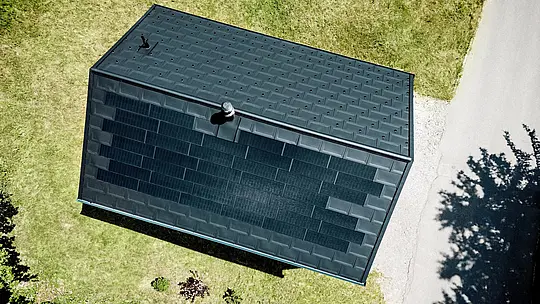 Spektakularna snimka dronom prikazuje PREFA solarni krov kuće Immi iz ptičje perspektive. Crne solarne krovne ploče gotovo se neprimjetno stapaju s krovnim pločama R.16 u boji P.10 Crna te oblikuju skladnu, kontinuiranu krovnu površinu. Integrirani aluminijski sustav objedinjuje održivu proizvodnju solarne energije, visoku otpornost i čistu arhitekturu, bez vizualno naknadno postavljene fotonaponske instalacije, prilagođene alpskom prostoru.