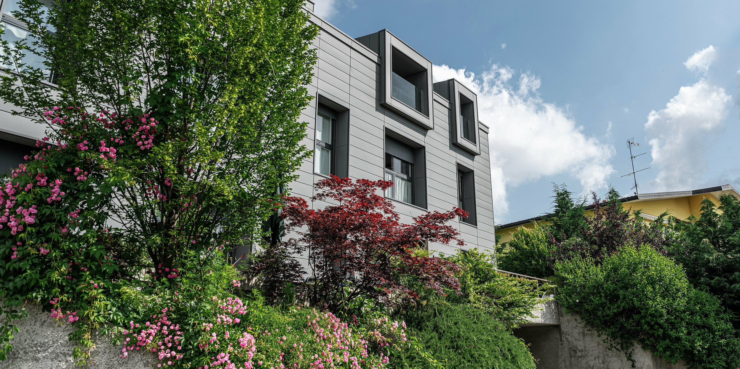 Moderne Wohnanlage in Faloppio, Italien, mit stilvoller Fassade aus PREFA Sidings. Die auskragenden Fenster bieten einen einzigartigen architektonischen Blickfang, während die umliegenden Pflanzen und Blumen die natürliche Schönheit des Objektes unterstreichen. Die langlebige Aluminiumverkleidung von PREFA sorgt für eine ästhetische und funktionelle Lösung bei jeder Witterung.