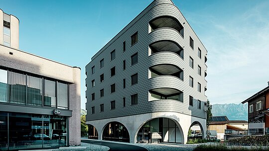 Ovaj moderni Bernerhaus u Buchsu oduševljava svojim upečatljivim zaobljenim, trokutastim oblikom zgrade i kontinuiranom fasadom od PREFA zidnih šindra u boji P.10 svijetlosivoj. Ljuskasta aluminijska površina naglašava nježne rubove i duboke zaobljene balkone, dok masivno betonsko postolje s elegantnim lukovima stvara zanimljiv kontrast. Usred male trgovačke ulice i na pozadini alpskog krajolika, lagana, otporna na vremenske uvjete i dugotrajna PREFA aluminijska fasada postaje arhitektonski vizualni naglasak: precizno je izvedena, gotovo bezdržavna i idealna za zahtjevne zaobljene oblike.