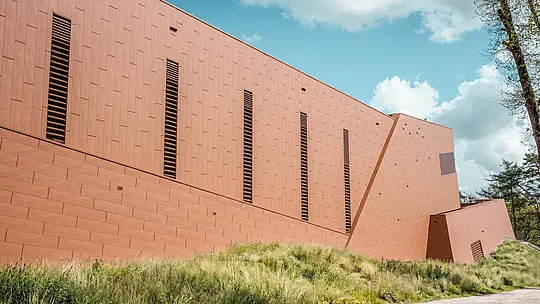 Upečatljiv bočni pogled na crpnu stanicu za pitku vodu WML u Lottumu s jasno strukturiranim, izduženim volumenom zgrade usred prirodnog krajolika livada. PREFA Siding.X aluminijska fasada u toplom izgledu hrđe strukturirana je ritmički postavljenim, vitkim lamelnim otvorima i stvara živahnu igru svjetla i sjene. Fino fasetirana površina, ovisno o perspektivi, djeluje dinamično i visokokvalitetno. Lagani, dugotrajni i potpuno reciklabilni PREFA aluminijski paneli jamče visoku otpornost na vremenske uvjete, postojanost boje te održiv i fasadni omotač zgrade koji zahtijeva minimalno održavanje.