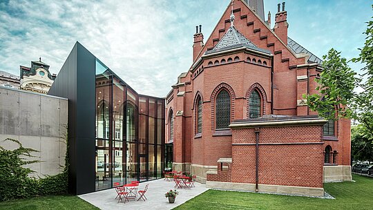 Slika prikazuje impresivnu novu zgradu koja povezuje povijesnu Crvenu crkvu s modernom istraživačkom knjižnicom u Olomoucu. Perspektiva prikazuje stražnji dio zgrade s pozivajućom terasom. Elegantna fasada od crnih PREFABOND aluminijsko-kompozitnih ploča postavlja upečatljiv, moderan naglasak i stilski kontrastira crvenom opečnom zidu crkve. Visoka preciznost montaže i upotreba dugovječnog materijala poput aluminija osiguravaju skladnu povezanost tradicije i inovacije. Ove napredne ploče nisu samo estetski dojmljive, već i izuzetno funkcionalne i dugovječne, čineći projekt remek-djelom moderne arhitekture.