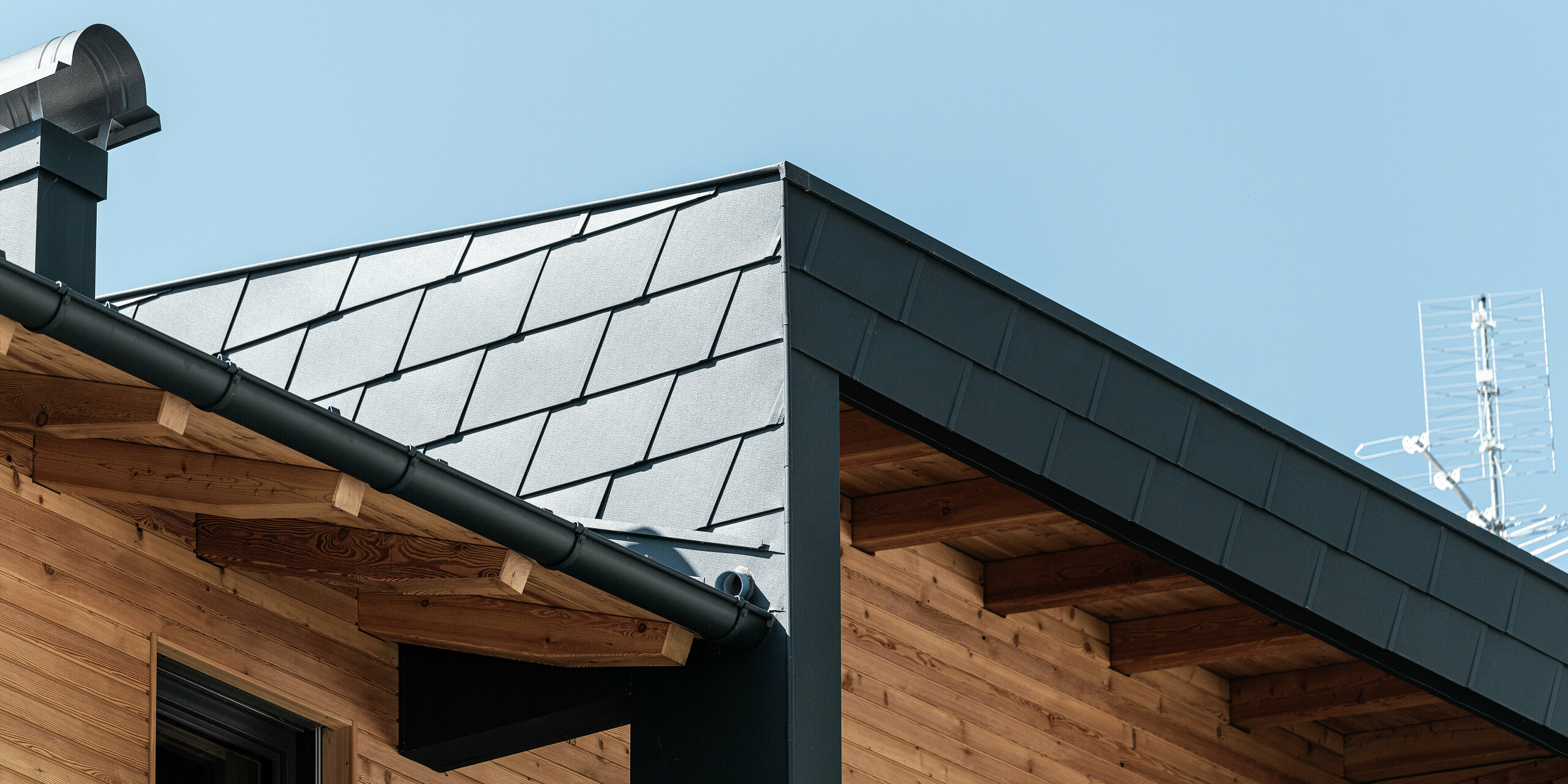 Detailansicht der Dachecke mit PREFA Dachschindeln DS.19 in der Farbe P.10 Anthrazit und der Fassadenverkleidung des überdachten Balkons. Die Kombination von Aluminium und Holz setzt ästhetische Akzente.