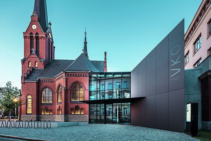 Slika prikazuje impresivnu igru između povijesne Crvene crkve i modernog novog objekta u Olomoucu. Crkva sa svojim karakterističnim gotičkim prozorima i upečatljivim tornjem čini povijesno središte ansambla, dok moderni prizid s elegantnom crnom fasadom od PREFABOND aluminijsko-kompozitnih ploča predstavlja suvremen, arhitektonski odgovor. Velike staklene površine omogućuju pogled u unutrašnjost nove zgrade, gdje se refleksijama u prozorima stvara zanimljiva interakcija između starog i novog. Precizna obrada PREFABOND ploča naglašava modernu estetiku, dok skladna integracija u povijesno okruženje impresionira i funkcionalno i vizualno.