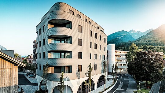 Kosi prikaz Bernerhausa u Buchsu s upečatljivom, trokutastom kubaturom i nježno zaobljenim kutovima. Kontinuirana fasada od PREFA zidnih šindra u P.10 svijetlosivoj boji obavija zgradu poput ljuskaste aluminijske kože i naglašava tekuće balkonske trake. Iznad masivnog betonskog postolja s elegantnim lukovima, lagana, otporna na vremenske uvjete PREFA aluminijska fasada djeluje posebno izražajno. Precizno postavljena, dugotrajna i gotovo bezdržavna, skladno se uklapa u alpsko okruženje i postavlja snažan arhitektonski naglasak.