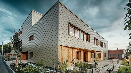 U nježnom večernjem ugođaju hibridna zgrada dječjeg vrtića u Alzenauu otkriva svoj puni arhitektonski dojam. Istaknuta fasada od brončano obojenih PREFA zidnih rombova 44 × 44 pruža zgradi elegantnu, vremenski otpornu „odoru“ s jasnom strukturom. U prizemlju vertikalne drvene obloge stvaraju prirodan, topao kontrast aluminiju te naglašavaju ulaz i zaštićene boravišne prostore. Reducirani, moderni oblikovni jezik skladno se uklapa u uređeni okoliš, u kojem klupe, biciklistička stalci i mlada stabla pozivaju na boravak. Ova slika dojmljivo prikazuje kako dugotrajni PREFA proizvodi doprinose stvaranju održivog, funkcionalnog, a istovremeno privlačnog obrazovnog objekta.