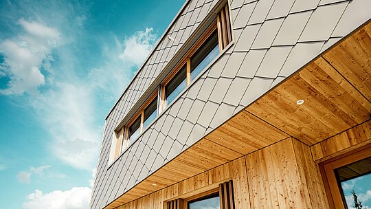 Detaljni prikaz dječjeg vrtića u Alzenauu otkriva preciznu izvedbu i visokokvalitetnu kombinaciju materijala – brončano obojenih PREFA zidnih rombova 44 × 44 i prirodnog drva. Dijagonalna struktura aluminijske fasade stvara upečatljiv uzorak „ljuski“, koji se skladno povezuje s vertikalnim drvenim lamelama na prozorima i fino obrađenim drvenim donjim rubovima. Snimka iz žablje perspektive naglašava točnost linija i majstorsku kvalitetu prednje ventilirane fasade. Ovaj detaljni prikaz ističe dizajnersku snagu i dugovječnost PREFA proizvoda – idealno za održive arhitektonske projekte koji na najvišoj razini spajaju estetiku, funkcionalnost i ekologiju.