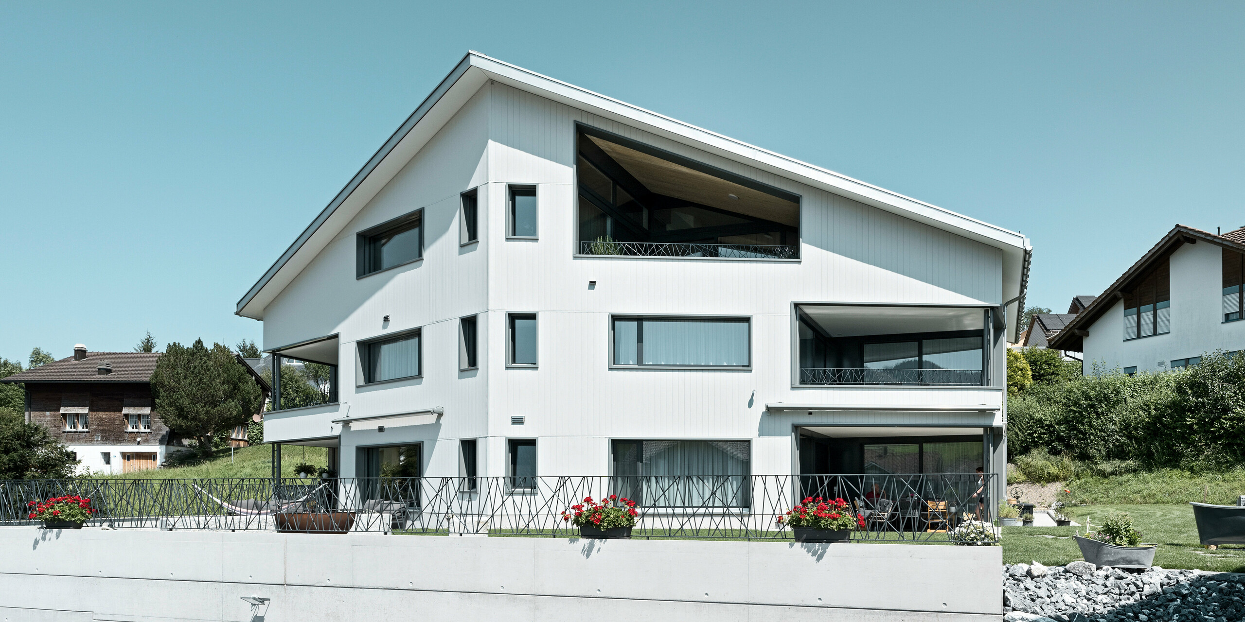 Das Bild zeigt ein Mehrfamilienhaus in Weissbad, Schweiz, das mit einer strahlend weißen PREFA Aluminiumfassade in P.10 Prefaweiß verkleidet ist. Die vertikale Ausrichtung der PREFA Sidings verleiht dem Gebäude eine elegante Textur und ein zeitgenössisches Flair. Die großzügigen Balkone und die weiten Fensteröffnungen sind harmonisch in das Fassadendesign integriert, wodurch eine ästhetische Symbiose aus Form und Funktion entsteht, die sowohl modern als auch einladend wirkt.