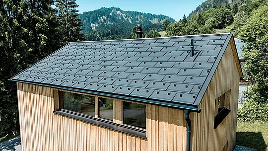 U svijetlom planinskom ljetu crni PREFA aluminijski krov kuće Immi dojmljivo dolazi do izražaja. Solarne krovne ploče integrirane u ravnini s krovom zajedno s krovnim pločama R.16 u boji P.10 Crna stvaraju mirnu i homogenu krovnu površinu. Lagani i robusni aluminijski sustav proizvodi električnu energiju iz sunca te pouzdano štiti drvenu kuću od zahtjevnih alpskih vremenskih uvjeta i velikih snježnih opterećenja.