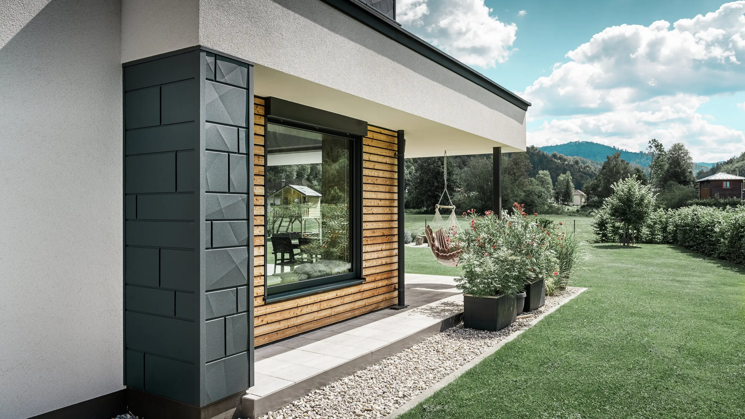 Djelomična snimka novoizgrađene obiteljske kuće. Na fasadi su korišteni različiti materijali. Bijela žbukana fasada je naglašena s PREFA aluminijskim Siding.X u antracit boji te horizontalnim drvenim panelima koji obuhvaćaju veliki prozor. Na terasi visi viseća stolica.