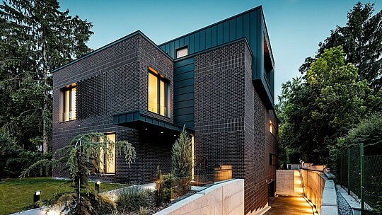 Snimka prikazuje modernu, jasno strukturiranu vanjsku vizuru budimpeštanske višegeneracijske vile u večernjem ambijentu. Tamnocrvenkasto-smeđe fasade od opeke susreću se s precizno izvedenim PREFALZ aluminijskim oblogama u boji P.10 antracit, koje svojim vertikalnim linijama stvaraju snažan arhitektonski naglasak. Toplo osvijetljeni prozorski otvori daju zgradi dubinu i atmosferu. Blago osvijetljen prilaz podzemnoj garaži elegantno prati volumen građevine i naglašava visokokvalitetnu, dugotrajnu i vremenski otpornu kombinaciju materijala klinkera i PREFA aluminija.