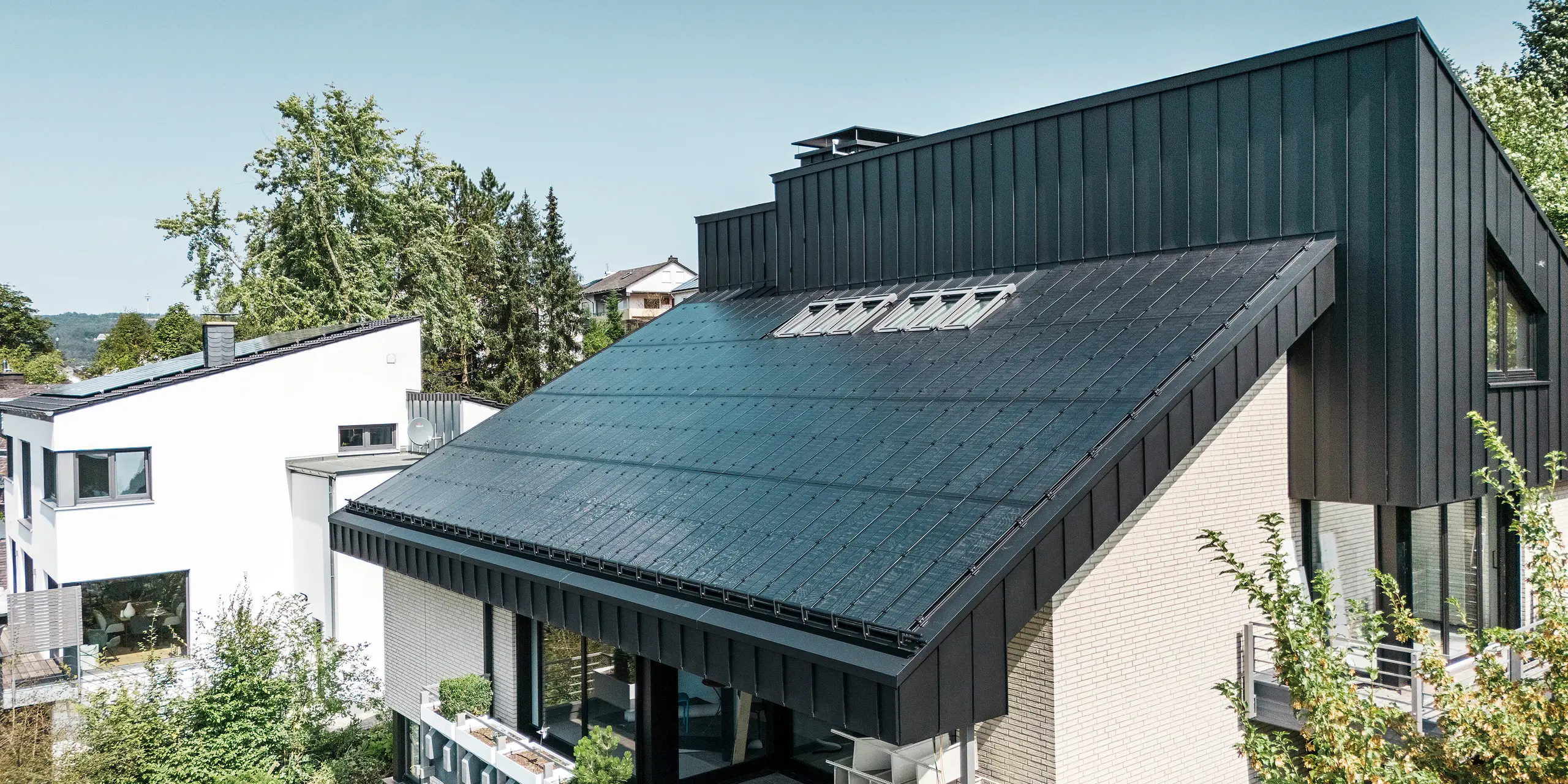Moderna arhitektura obiteljske kuće u Meschedeu, Njemačka, s PREFALZ aluminijskim krovom u P.10 crnoj boji i integriranim staklo/staklo PV modulima. Pomaknuti krov spaja bezvremenski dizajn s održivom fotonaponskom tehnologijom. Inovativni PREFALZ solarni moduli pružaju snagu od 18 kWp. Bezokvirni moduli i elegantna aluminijska obloga krova i fasade čine estetsku i funkcionalnu cjelinu koja se savršeno uklapa u zeleno stambeno okruženje.