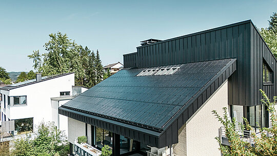 Moderna arhitektura obiteljske kuće u Meschedeu, Njemačka, s PREFALZ aluminijskim krovom u P.10 crnoj boji i integriranim staklo/staklo PV modulima. Pomaknuti krov spaja bezvremenski dizajn s održivom fotonaponskom tehnologijom. Inovativni PREFALZ solarni moduli pružaju snagu od 18 kWp. Bezokvirni moduli i elegantna aluminijska obloga krova i fasade čine estetsku i funkcionalnu cjelinu koja se savršeno uklapa u zeleno stambeno okruženje.