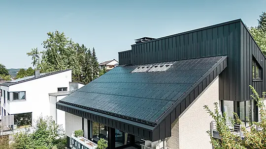 Moderna arhitektura obiteljske kuće u Meschedeu, Njemačka, s PREFALZ aluminijskim krovom u P.10 crnoj boji i integriranim staklo/staklo PV modulima. Pomaknuti krov spaja bezvremenski dizajn s održivom fotonaponskom tehnologijom. Inovativni PREFALZ solarni moduli pružaju snagu od 18 kWp. Bezokvirni moduli i elegantna aluminijska obloga krova i fasade čine estetsku i funkcionalnu cjelinu koja se savršeno uklapa u zeleno stambeno okruženje.