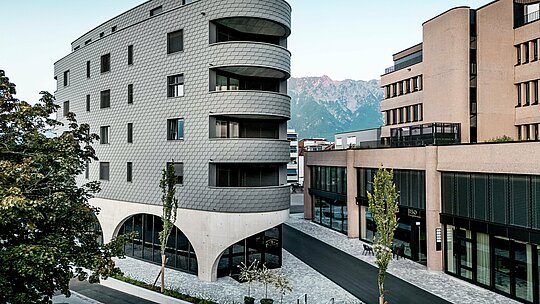 Pogled na Bernerhaus u Buchsu s upečatljivim trokutastim oblikom i nježno zaobljenim balkonima na pozadini alpskog krajolika. Kontinuirana fasada od PREFA zidnih šindra u P.10 svijetlosivoj boji obavija zgradu poput ljuskaste aluminijske kože i stvara živopisnu igru svjetla i sjene. Iznad masivnog betonskog postolja s elegantnim lukovima, lagana, otporna na vremenske uvjete PREFA aluminijska fasada djeluje posebno precizno, dugotrajno i gotovo bezdržavno – arhitektonski privlačan element u urbanom okruženju.